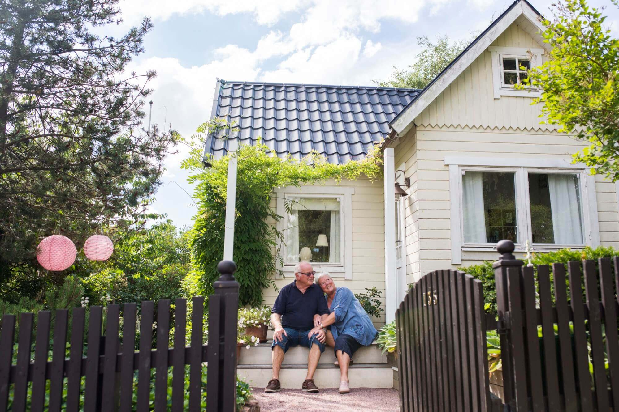 An elderly couple sits at the entrance to their bright wooden cottage with a blue roof and greenery all around after painting the facade.